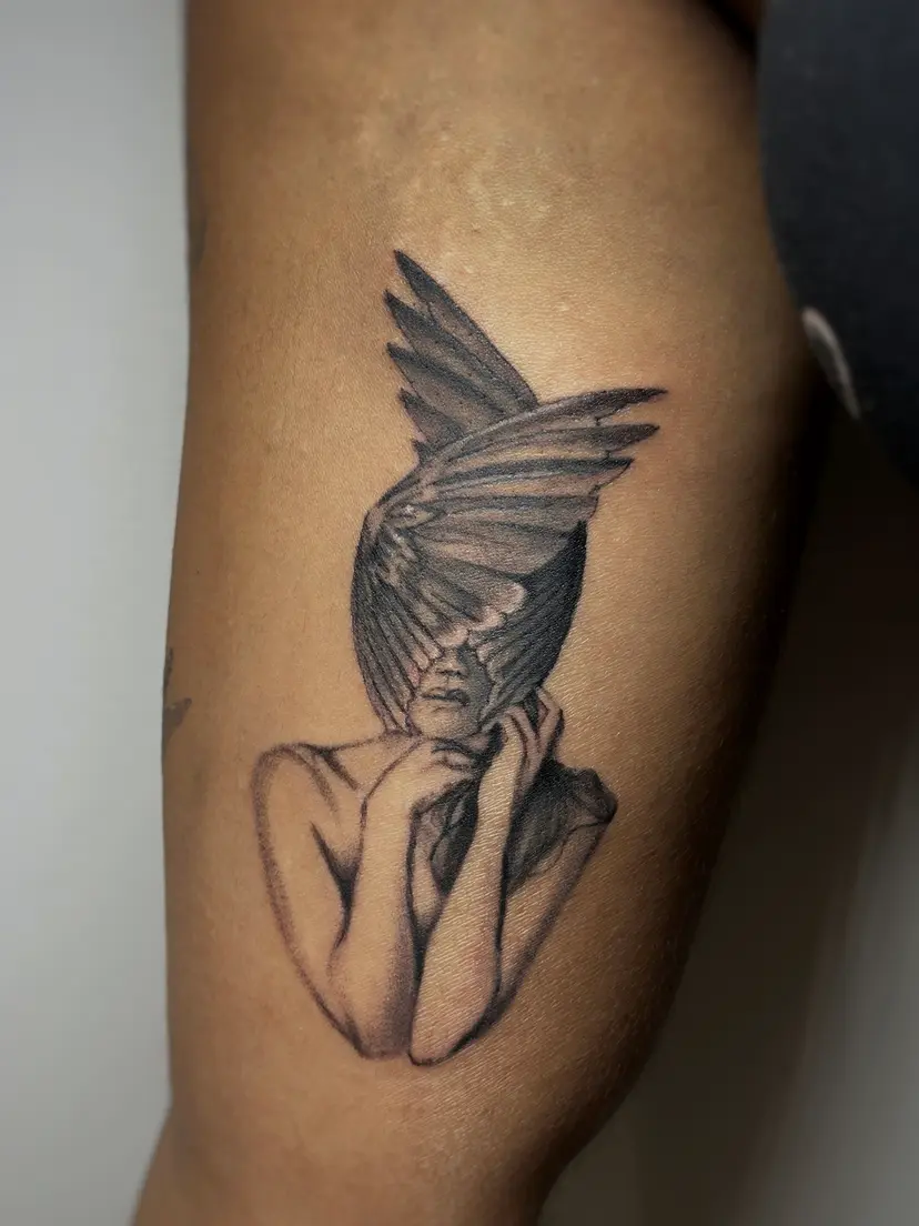 Black and grey tattoo of a woman whose face is hidden by wings, on an arm