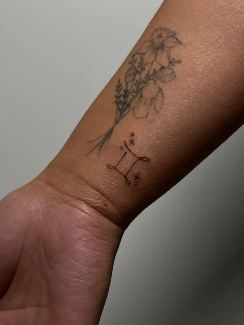 Fine-line bouquet and Gemini sign tattoo on a forearm